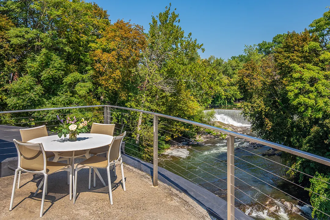 Outdoor terrace with table and chairs overlooking a river and waterfall surrounded by trees.