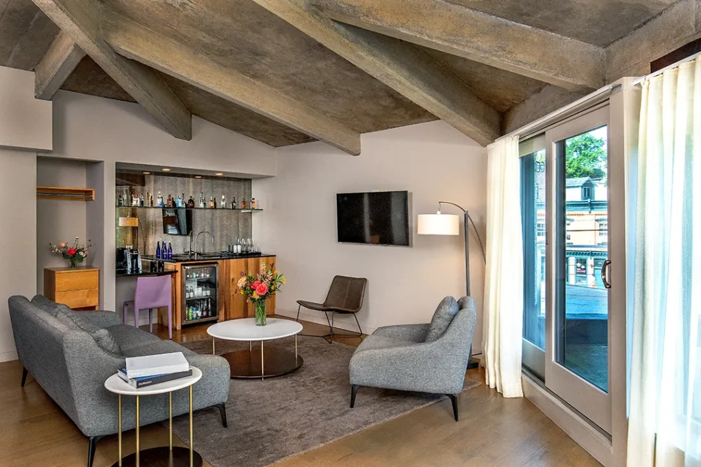 Modern living room with exposed beams, seating area, minibar, and large glass doors.