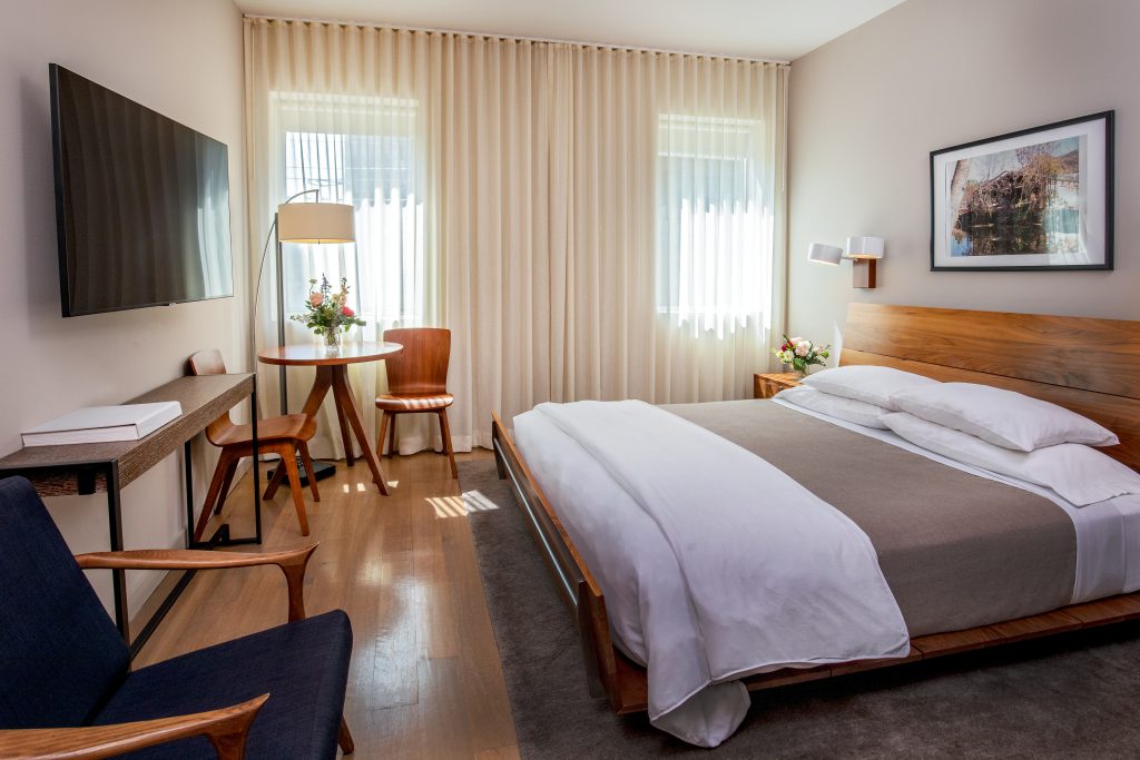Modern hotel room with wooden bed, desk, tv, and soft daylight through sheer curtains.