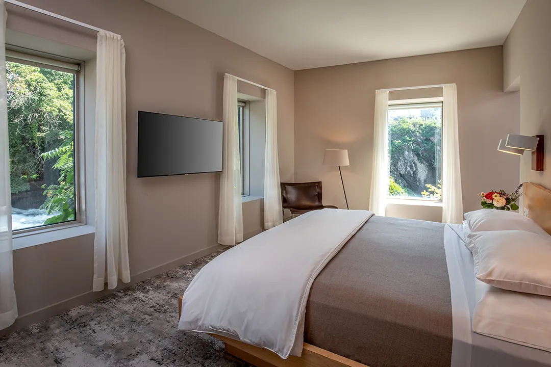Minimalist hotel bedroom with large bed, soft lighting, and river-view windows.
