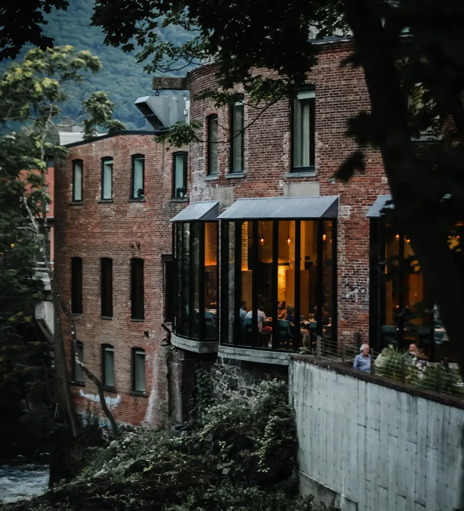 Brick riverside building with large windows glowing at dusk, people dining inside.