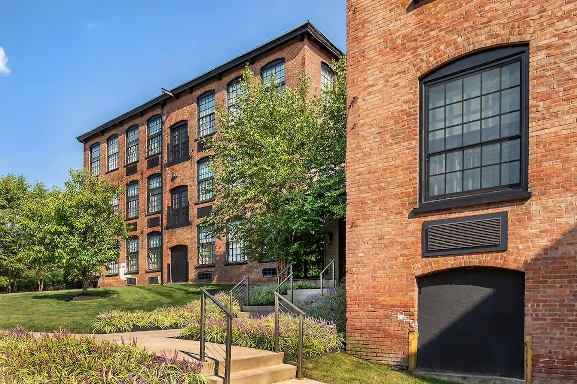 Brick mill buildings with black-framed windows, landscaped paths, and trees under a clear blue sky.